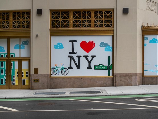 A storefront window is covered by a poster featuring the phrase "I ❤️ NY" with a green bicycle illustration and a street sign for "Fifth Ave." The backdrop includes simplistic graphics of buildings and clouds