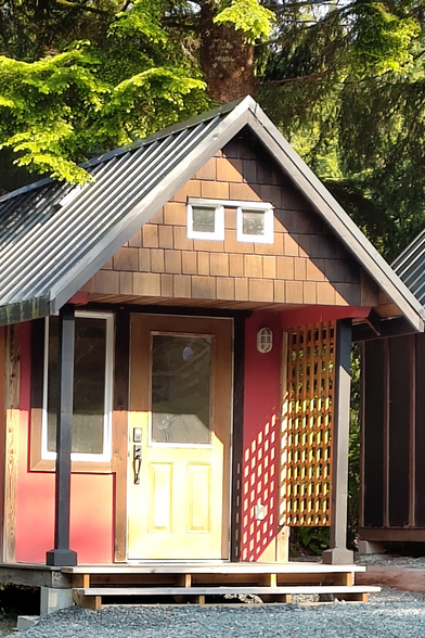 A one room camping pod cabin in sheathing ply and cedar shingles under a felt roof. The gable end has a porch overhanging the yellow door. Two fixed lights like eyes above. A trellis casts shadow from the low sun