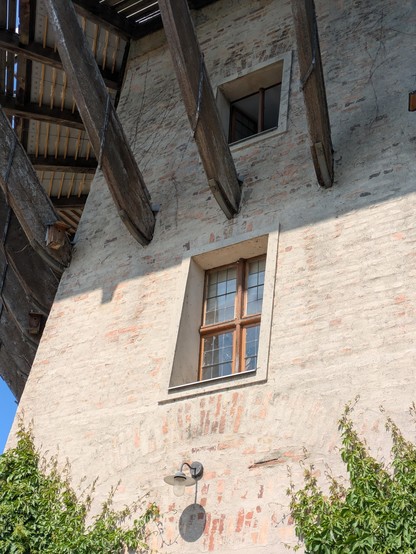 Von schräg unten gesehen: Teilansicht einer alten Windmühle mit einem Fenster im Zentrum einer nur dünn verputzten Ziegelwand. Es hat einen Holzrahmen, blauer Himmel spiegelt sich im Glas. Unter dem Fenster befindet sich eine metallene Lampe, die Tür, die sie beleuchtet, ist nicht mehr im Bild. Von rechts und links unten erobert wilder Wein die Wand. Über dem Fenster ragen kräftige Balken schräg nach oben und tragen eine breite umlaufende hölzerne Galerie. In deren Schatten liegt noch ein kleineres Fenster.