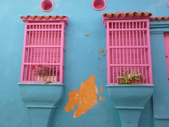 Blaue Hausfront mit knallrosa Fenstern, die rosa vergittert sind und auch von innen rosa Fensterläden haben.