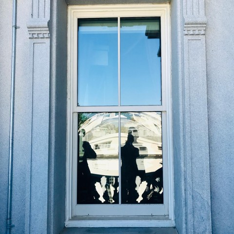 A white-framed sash window set in a gray stone facade with decorative columns. The window has two panes - the upper pane shows a blue sky reflection, while the lower pane reflects the curved architecture of the White House's South Portico with its distinctive columns and rounded structure. Two silhouetted figures are visible in the foreground of the reflection.