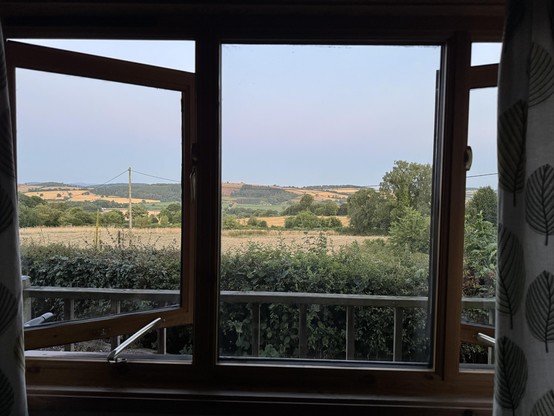 A wooden window is open, revealing a scenic view of rolling hills, fields, and trees under a light sky. A hedgerow frames the foreground, adding to the rural ambiance.
