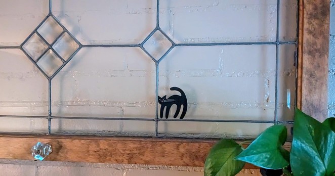 A leaded glass window hangs in front of a wall with a decal of a black cat in one of the panes.
