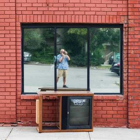 The photographer's reflection is seen in a large window set in a red brick wall. An old tv and stand lie sideways on the sidewalk below the window.