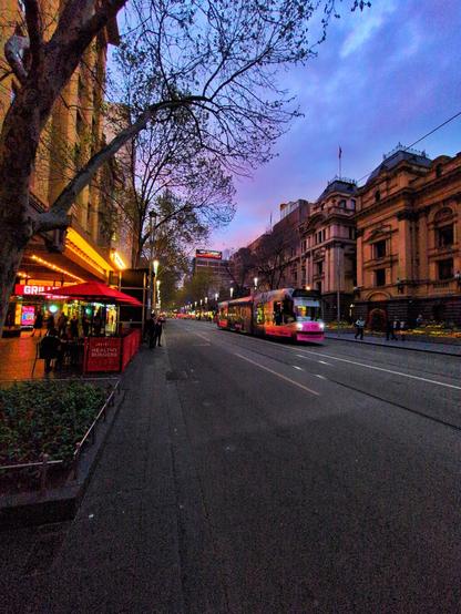 A pink tram rides past the sandstone Town Hall, on the left. A small outdoor seating area with red umbrellas is on the left. The sky is light blue and purple.