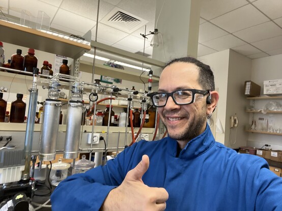 I middle aged whiteish-looking man wearing safety glasses and a flame resistant lab coat gives a thumbs up, standing in front of a Schlenk line