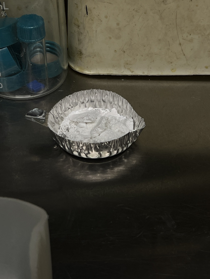 White powdered in a crimped aluminum weigh boat. Photo taken through the window of an inert atmosphere glove box