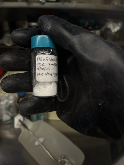 A bulky black rubber glove holding a 20 mL glass vial with a green cap containing a white powder. Vial labeled “(tBuSalAmEE)Al  YDG-3-003  4/10/23  tare w/cap: 18.200 g”