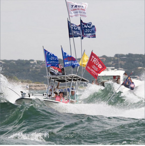 A photo of a boat with several large MAGA and Trump 2020 flags flying from a small boat being swamped by waves created by bigger boats in the 'Trump Armada' on Lake Travis, Texas,  This and several other boats sunk. No morons died that day.
