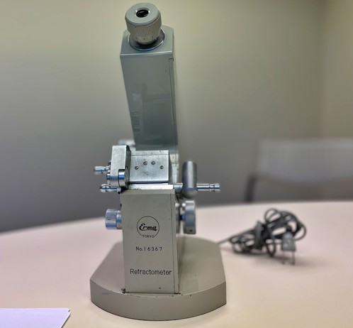 .  A metal instrument with an eyepiece and several dials on the right and left and an additional metal cover that swings open for sample disposition.  In the back, there is a cord to plug in a light source. The instrument has the incription, "Erma Tokyo No. 16367.  Refractometer"