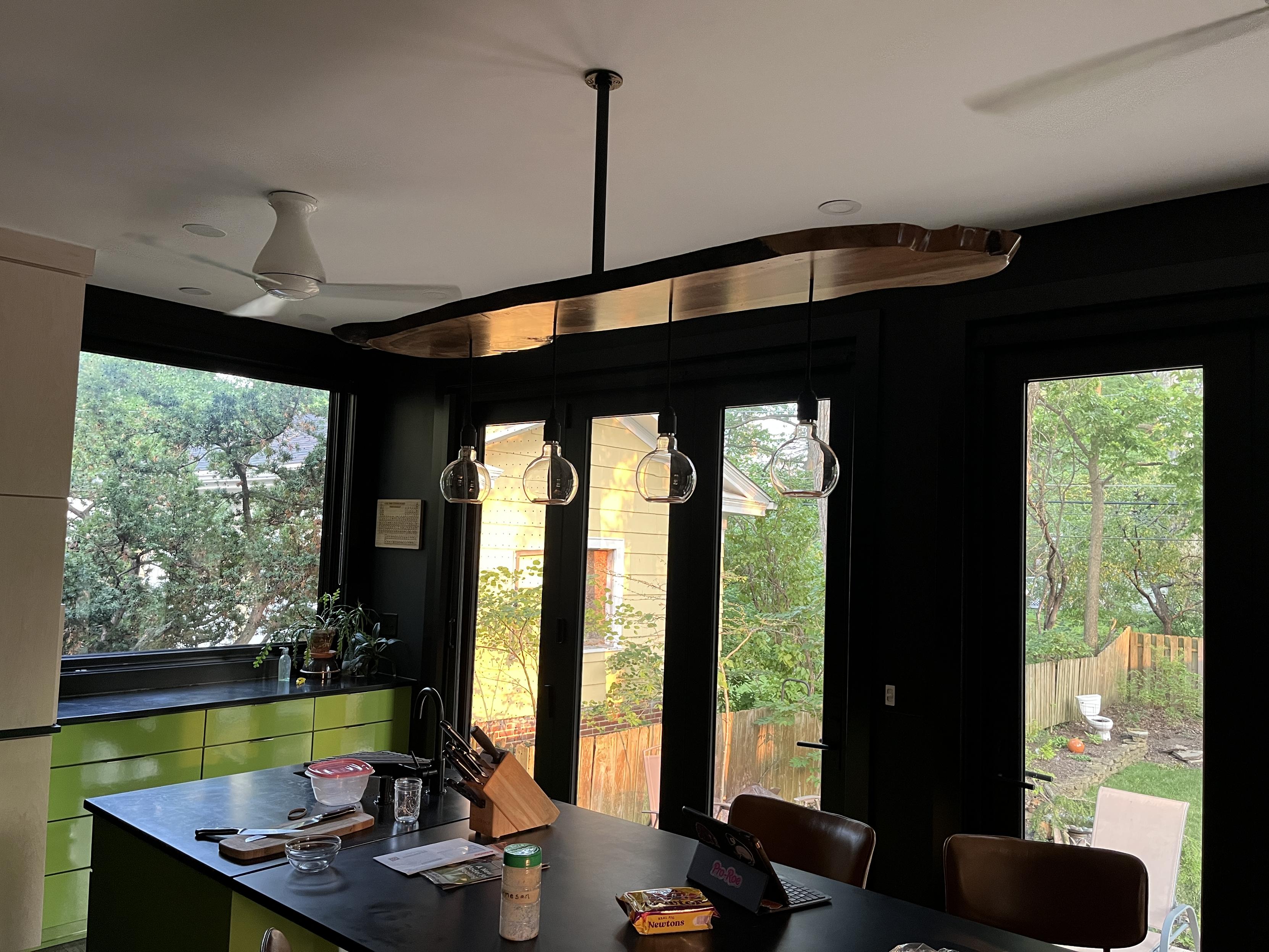 A long slab of wood hangs from a single black iron pipe. At the top of the pipe is a flange with screw heads visible. Four large partially open globes hang below it on cloth cables. They are set in simple black sockets. The finish warmly reflect light that shines in from the windows in the background. The counters and table are black and the cabinets are bright green or white maple.
