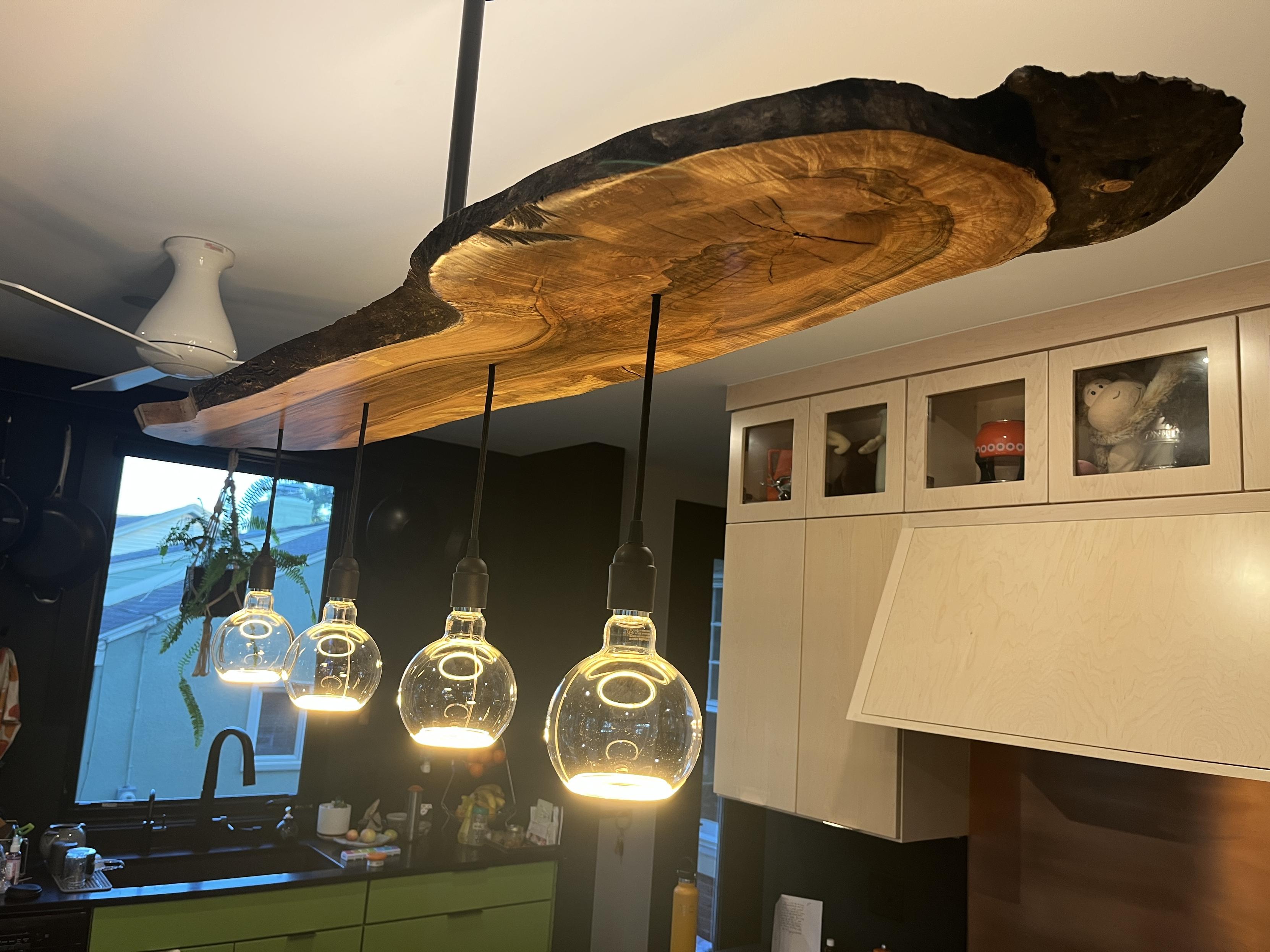 A long live-edge slab of wood hangs from a single black iron pipe that extends out of frame. Four large partially open globes set in simple black sockets hang below it on cloth cables, glowing from their lower rims. They lights the highly polished wood surface above, which is rich in reds, golds, and browns. The ceiling above the fixture is also lighted. In the background are white maple cabinets, a range hood, and a copper backsplash.