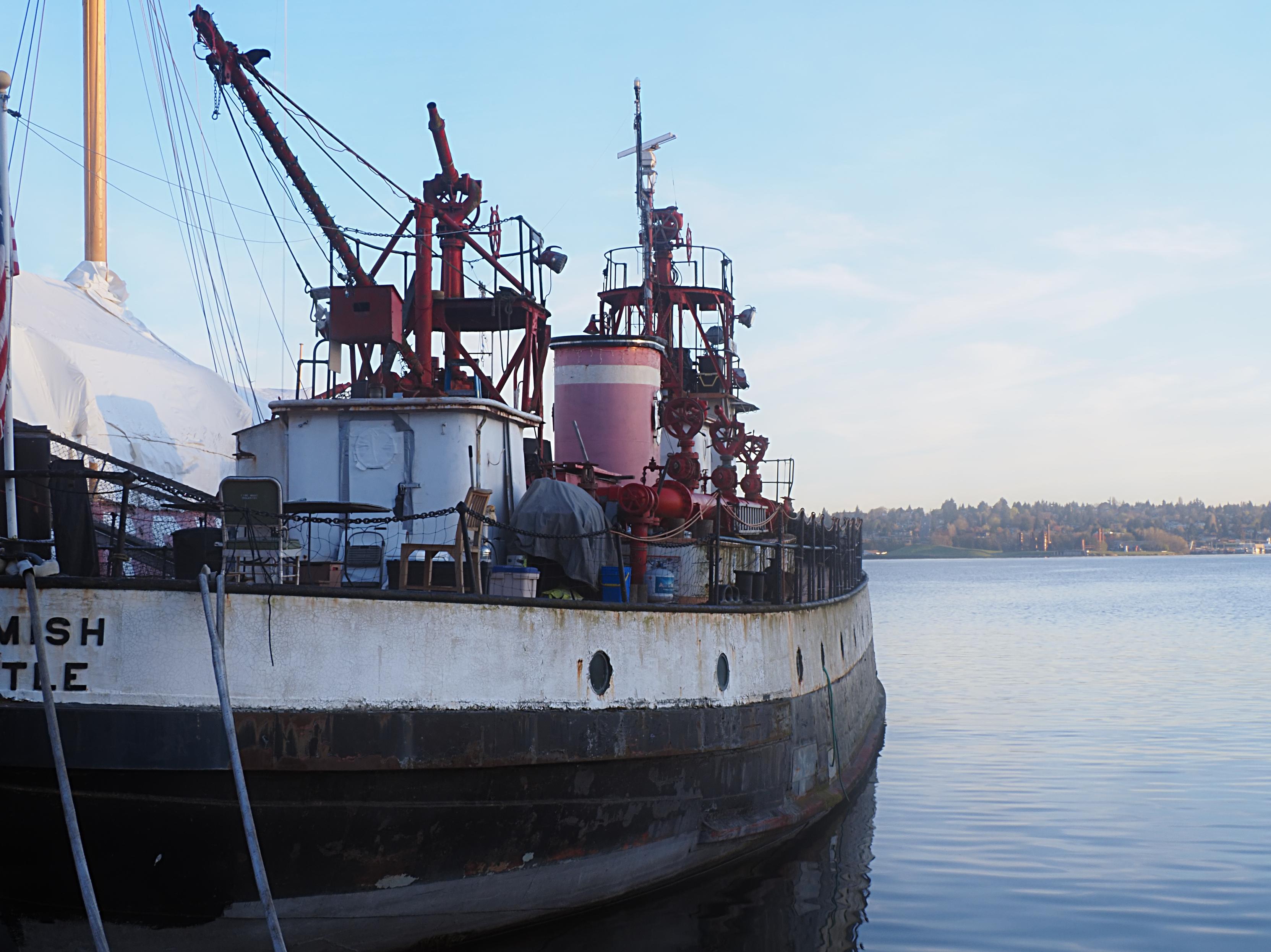 A boat with a black hull and red equipment in a state of disrepair on a lake; the morning sun behind.  The historic Fireboat Duwamish at Lake Union Park in Seattle.  There is a family of raccoons on the deck near the fencing.