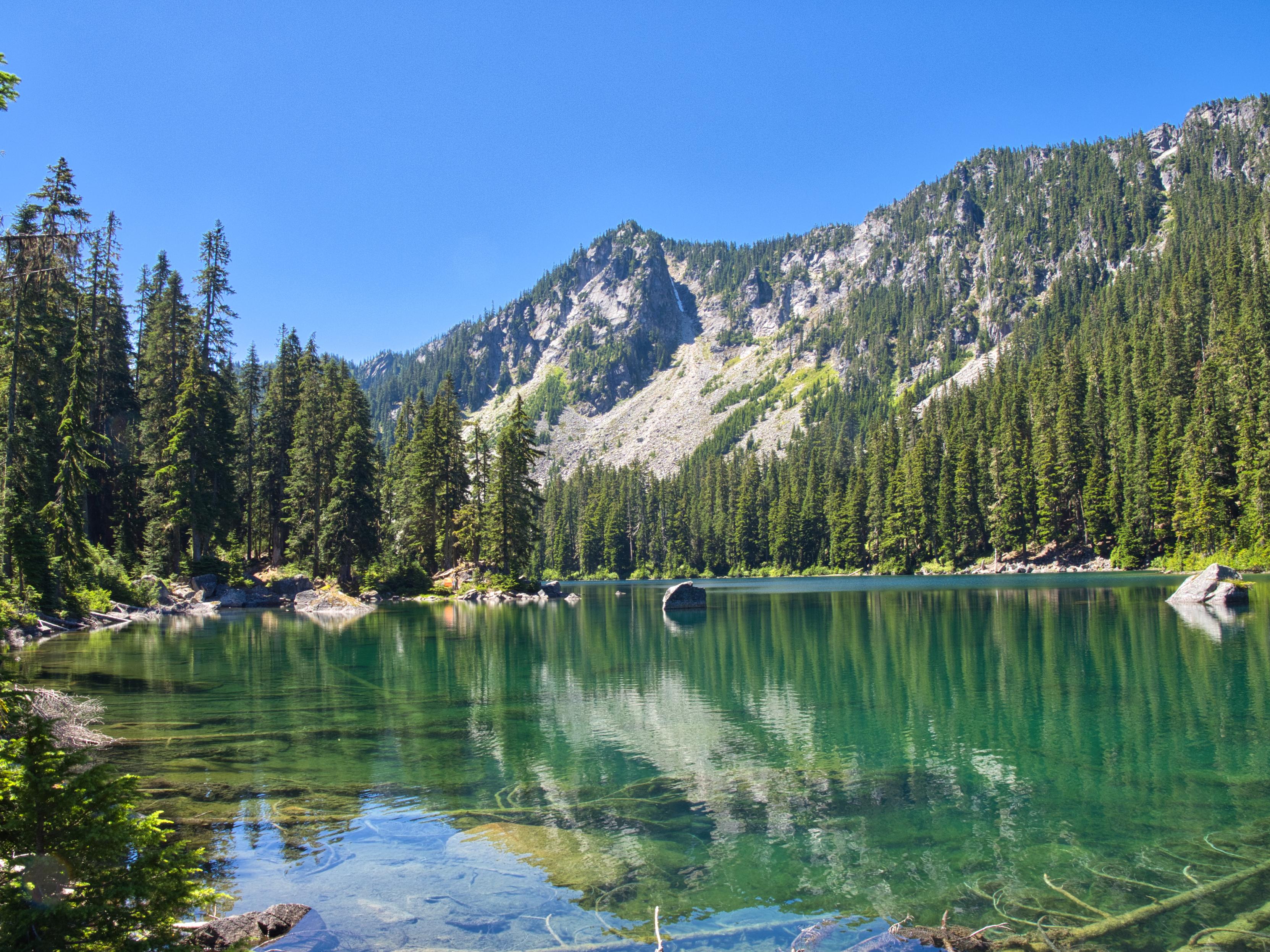 A lake of clear green water reflecting the trees and white rocks of the mountain above the far shore.